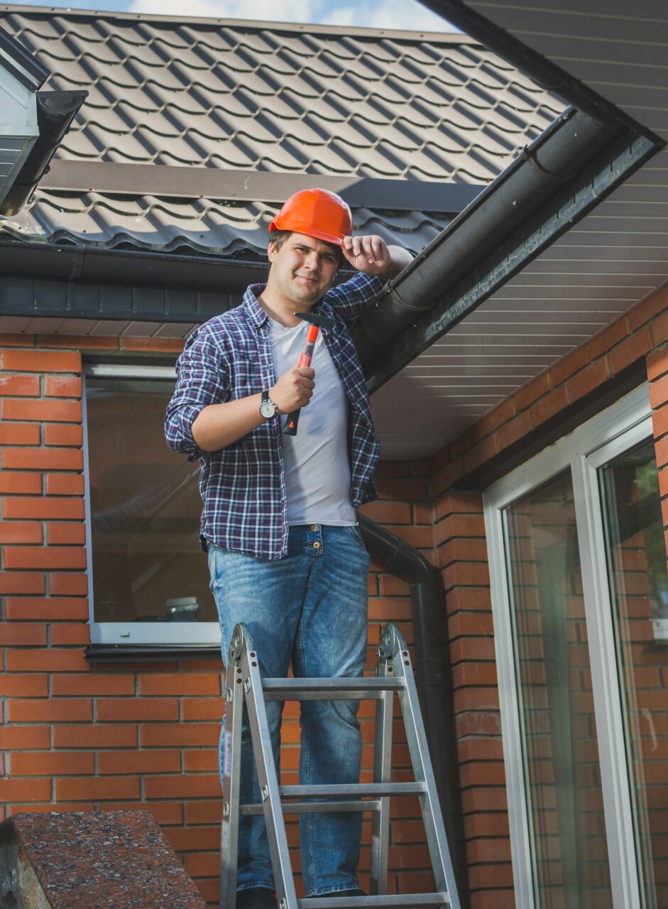 smiling carpenter posing with hammer on step ladder under the roof